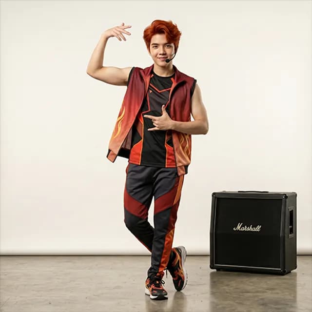 Dance teacher with red hair in a flame-pattern vest and headset mic striking a pose beside a Marshall amp
